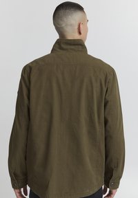 Jachetă verde măslinie cu guler ridicat și manșete cu nasturi, având un material texturat și o croială lejeră, văzută din spate.