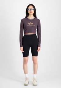 Fialový krátky top s dlhými rukávmi a zlatým logom, skombinovaný s čiernymi rebrovanými bikerskými šortkami a béžovými teniskami, doplnený o reflexné slnečné okuliare.