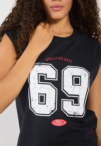 Svakjort svart uten ermer med store hvite sprukne tall "69" og rød tekst "ATHLETICS DEPT." med en liten rød "NYC 98" logo.