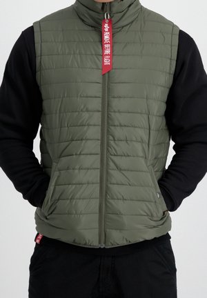 Man wearing olive green quilted vest with front zipper and red "Remove Before Flight" tag, paired with black long-sleeve shirt and pants.