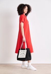 Röd kortärmad klänning med avslappnad passform. Svart vävd handväska med vita accenter och logotyp. Vita sneakers med ankelsockor.