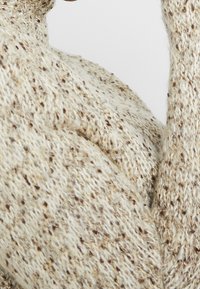 Sweter dziergany w kolorze kremowym z brązowymi flekami, z teksturowaną tkaniną i luźnym krojem, podkreślający miękkość i ciepło materiału.