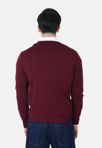 Uomo di spalle che indossa un maglione bordeaux sopra una camicia bianca e jeans blu su uno sfondo chiaro e semplice.