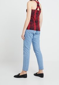 Bluzka w czerwono-czarny wzór wężowy z krzyżowym tyłem, zestawiona z jasnoniebieskimi dżinsami i czarnymi butami na slip-on, stojąca na neutralnym tle.