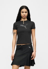 Γυναίκα με μακριά σκούρα μαλλιά, φορώντας μαύρο τοπ Puma με φερμουάρ μπροστά και μαύρη φούστα Puma, στέκεται μπροστά σε απλό λευκό φόντο.