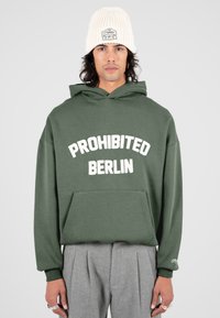 Hombre con una sudadera verde que tiene el texto "PROHIBIDO BERLÍN", pantalones grises con pliegues, gorro blanco de punto y varios anillos en los dedos.