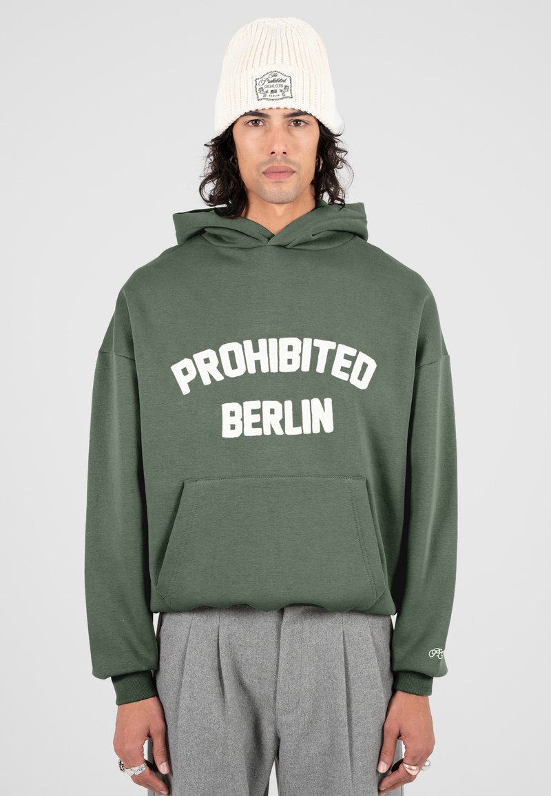 Hombre con una sudadera verde que tiene el texto "PROHIBIDO BERLÍN", pantalones grises con pliegues, gorro blanco de punto y varios anillos en los dedos.