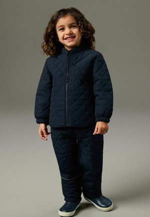 Uśmiechnięte dziecko o kręconych włosach, ubrane w granatową, pikowaną kurtkę i spodnie, stojące na jednolitym szarym tle, ubrane w ciemne buty.