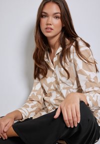 Camicia button-up beige e bianca con un motivo a forme astratte e colletto, abbinata a pantaloni neri a vestibilità ampia.