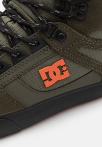 Maslinasto zelena visoka obuća napravljena od antilopa i tkanine, s crnim gumeni potplatom i narančastim DC logotipom sa strane. Crne i sive vezice.