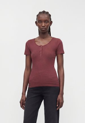 Jeune femme aux cheveux tressés porte un haut côtelé bordeaux ajusté à manches courtes avec des boutons et un pantalon noir, sur un fond uni.