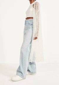 Kvinna som bär ljusblå vida jeans, vit crop top, vita sneakers och en lång genomskinlig vit blomsterbroderad cardigan.