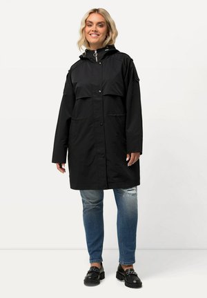 Black waterproof parka jacket with a hood, two front pockets, and snap button closure, paired with distressed blue jeans and black loafers.