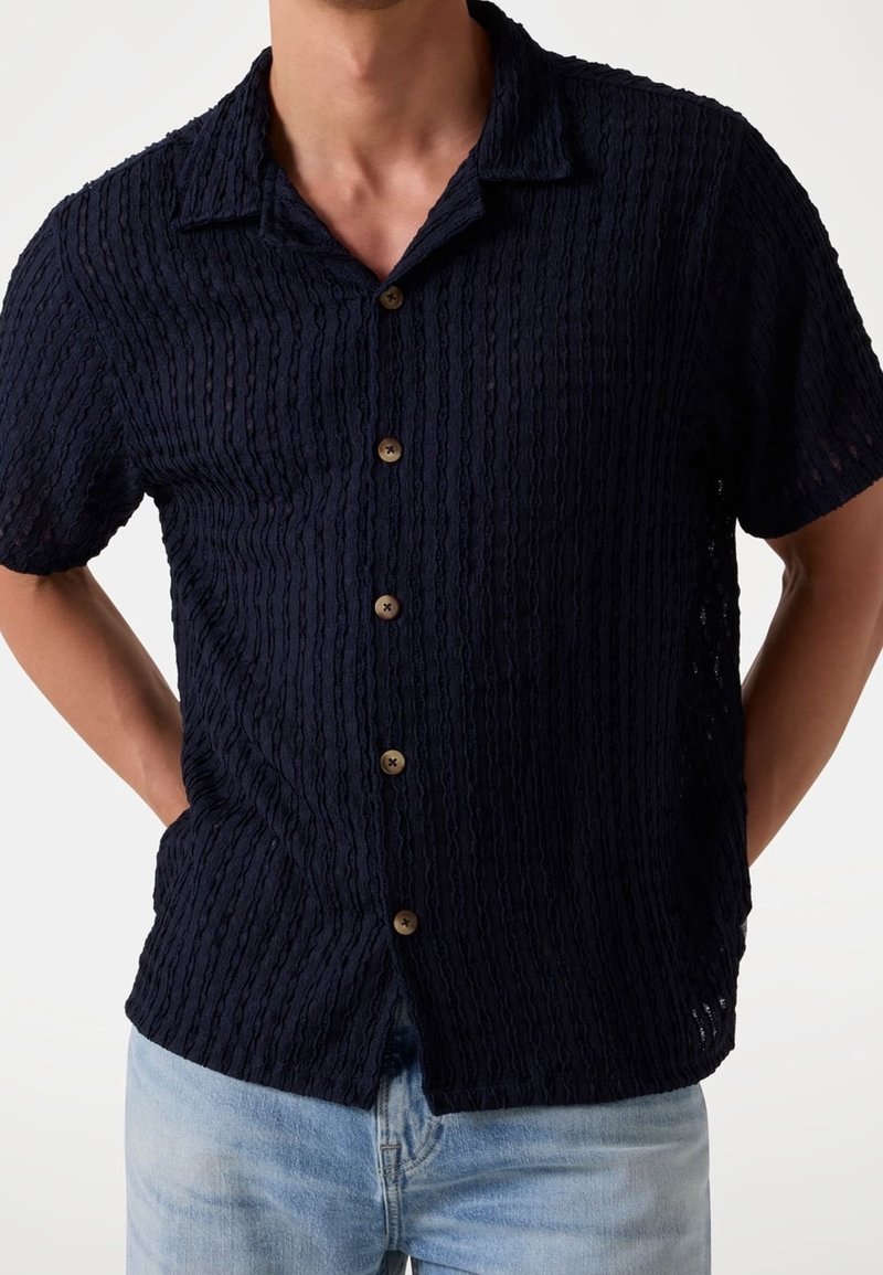 Homme portant une chemise à manches courtes bleu marine texturée à boutons, avec un col décontracté et un jean bleu clair, mains derrière le dos.