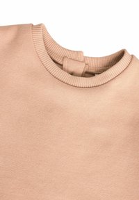 Ljust beige tröja med en ribbad rund halsringning och en mjuk, slät textur. En anmärkningsvärd detalj är en liten ögla vid nacke bak.