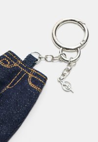 Un porte-clés en métal argenté avec une chaîne attachée à un tissu en denim bleu foncé avec des coutures jaunes sur fond blanc.