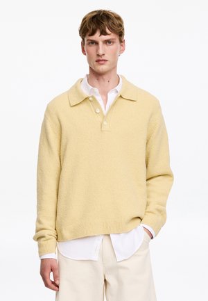 Ung mand iført en lys gul tekstureret sweater over en hvid knap-op skjorte og cremefarvede bukser mod en ensfarvet hvid baggrund.