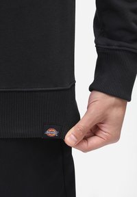 Černá mikina se žebrovanými manžetami a lemem, s viditelnou etiketou v dolním levém rohu, na které je logo "Dickies".