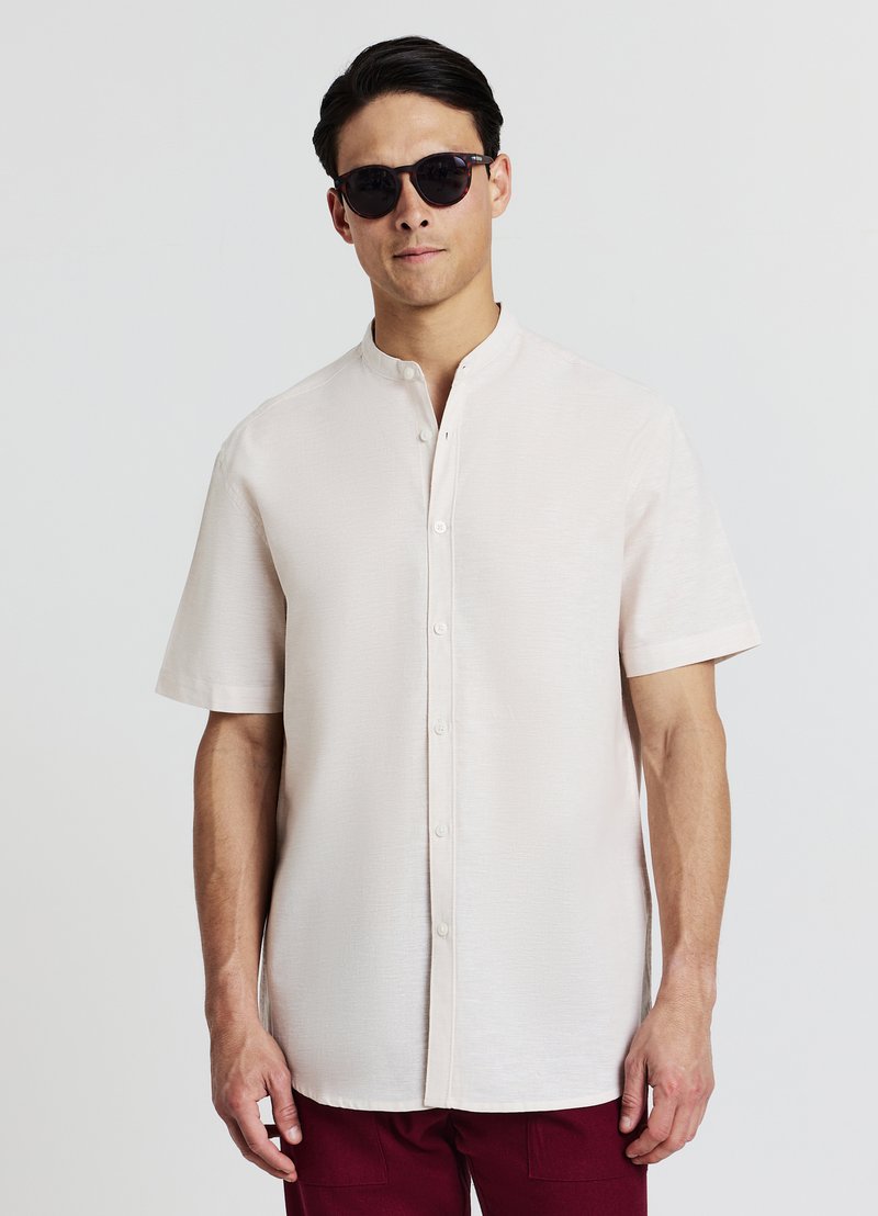 Chemise beige clair à manches courtes avec boutons et col mao, en tissu texturé avec un éclat subtil. Portée avec des lunettes de soleil foncées.