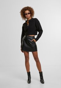 Zwarte cropped cardigan met knopen, gecombineerd met een zwarte leren minirok met hoge taille en zwarte enkelboots. Geweven krullend haar.