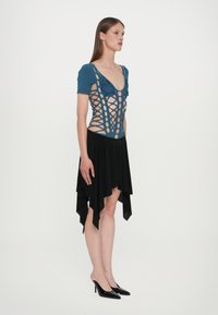 Τιρκουάζ ολόσωμο ρούχο με σχέδιο κοπής και κοντά μανίκια, συνδυασμένο με μαύρη ασύμμετρη φούστα. Το μοντέλο φοράει μαύρες ψηλοτάκουνες γόβες.