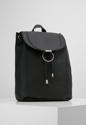 Sac à dos en cuir noir avec un rabat avant et un détail d'anneau argenté, posé sur une surface blanche contre un fond gris uni.
