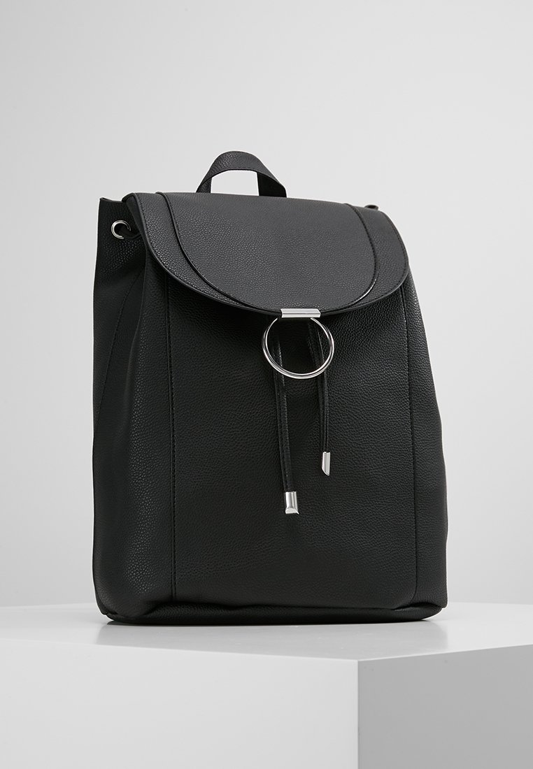 Sac à dos en cuir noir avec un rabat avant et un détail d'anneau argenté, posé sur une surface blanche contre un fond gris uni.