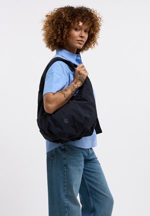 Vrouw met krullend haar die een blauwe blouse en spijkerbroek draagt, met een grote zwarte schoudertas met het "GOT BAG"-logo, toont een handtattoo.