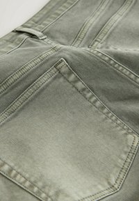 Gros plan sur un tissu en denim vert délavé montrant la poche arrière cousue et la boucle de ceinture du jean.