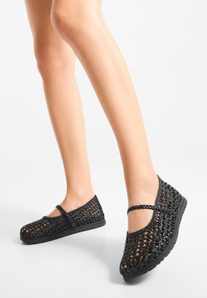 Musta kudottu tasapohjainen kenkä, jossa on suljettu kärki ja Mary Jane -remmi. Verkkokuviointi mahdollistaa hengittävyyden, ja pohja on tasainen kumia.