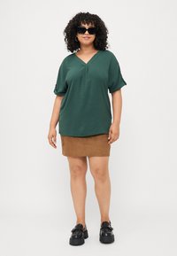 Ciemnozielona, teksturowana bluzka typu V-neck z krótkimi, zrolowanymi rękawami, w połączeniu z brązową, zamszową mini spódnicą oraz czarnymi platformowymi mokasynami.