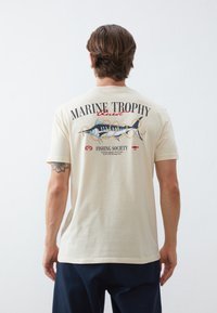 Cotton t-shirt, cream color, featuring a large graphic design of a fish and text on the back. Short sleeves, casual fit, plain front.