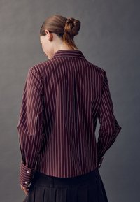 Bordó csíkos ing nyakán gallérral, hosszú ujjal, fehér függőleges vonalakkal és gombos mandzsettákkal. Testhezálló szabású, hátul varrással díszítve.