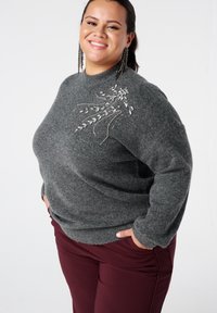 Grijze gebreide trui met een hoge halslijn, met een bloemenpatroon van kralen aan de linkerkant. Gecombineerd met bordeauxrode broek.