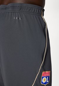 Grå idrætsshort med en lynlås lomme, prydet med et logo fra Olympique Lyonnais og "AEROREADY" branding på taljen.