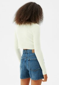 Osoba s kudrnatými vlasy, oblečená v bílé žebrované krátké mikině s dlouhým rukávem a vysokými modrými džínovými šortkami, stojící zády k bílé pozadí.