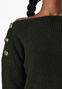 Tamnozeleni pleteni pulover s off-shoulder dizajnom i četiri velika, okrugla dugmeta na lijevom ramenu. Mekana tekstura s debelim uzorkom pletenja.