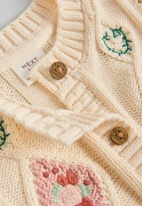 Creme strikket cardigan med blomsterbroderi, ribbede kraver og to brune knapper med logo. Blød tekstur og varme designelementer.