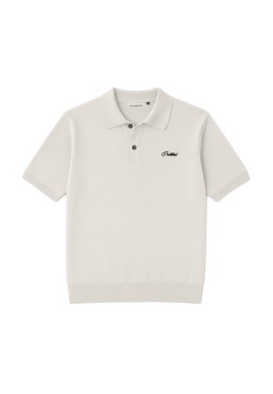 Ung mand iført en hvid polo med broderet tekst og en beige og sort trucker-kasket, der står foran en lysegrå baggrund.