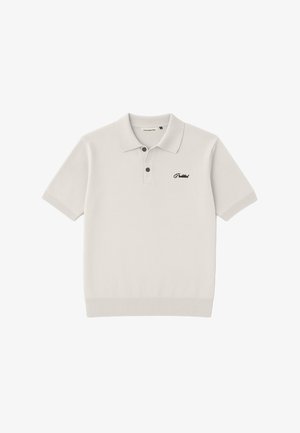 Młody mężczyzna w białej koszulce polo z wyszywanym napisem i beżowo-czarnej czapce z daszkiem stoi na tle jasnoszarym.