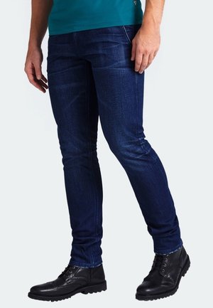Homme portant un jean slim bleu foncé, des bottes en cuir noires à lacets et une chemise turquoise, debout avec les mains détendues le long du corps.