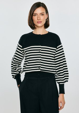 Kobieta z krótkimi brązowymi włosami, ubrana w czarno-biały kardigan w poziome paski i czarne spodnie z wysokim stanem, stojąca z jedną ręką w kieszeni.