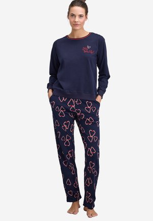 Vrouw met marineblauwe top met lange mouwen met tekst "Stay Beautiful" en bijpassende marineblauwe broek met rode en witte hartjes, staand op blote voeten.