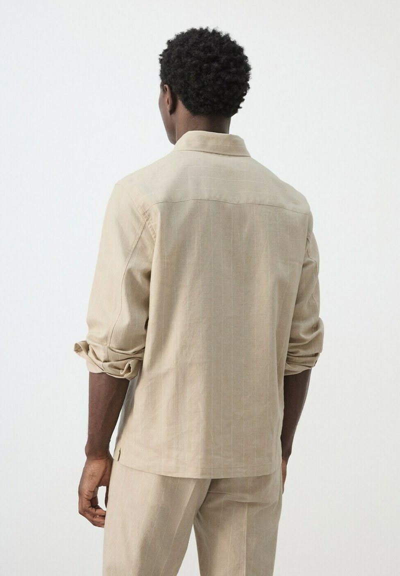 Homme portant une chemise beige à manches longues avec les poignets retroussés et un pantalon assorti, debout de dos sur un fond blanc uni.