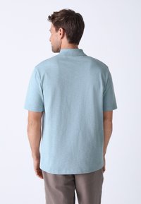 Polo bleu clair, manches courtes, tissu texturé et ourlet droit. Porté avec un pantalon beige, affichant un design simple.