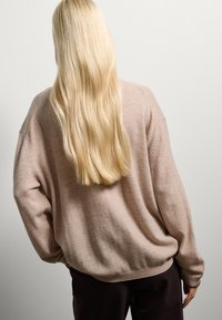 Pull en maille beige clair, à coupe ample et épaules tombantes, associé à un pantalon sombre. Texture lisse et cheveux longs et raides visibles.