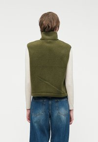 Zeleni flis brezrokavnik z visokim ovratnikom, ki ima mehko teksturo in skrajšano obliko, nosi pa ga čez svetlo obarvano dolgotrajno majico.