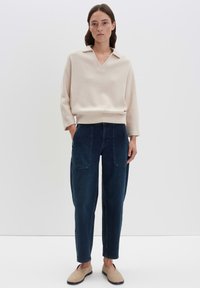 Frau steht da und trägt einen cremefarbenen, langärmeligen V-Ausschnitt-Pullover, dunkelblaue, hochtaillierte Hosen mit Fronttaschen und beige Slipper.