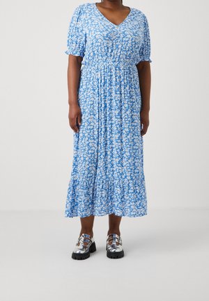 Femme portant une robe midi florale bleue avec des manches courtes bouffantes et des chaussures argentées épaisses sur un fond clair uni.