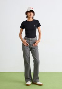 Sort tætsiddende t-shirt, grå højtaljede jeans og hvide sneakers med brune såler. Bærer en hvid kasket med rød tekst. Grøn gulvbaggrund.